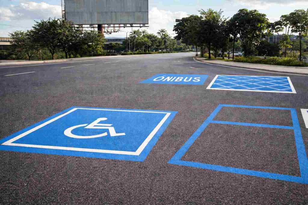 Imagem realista de uma via urbana moderna exibindo exemplos de sinalização horizontal azul conforme aplicada no trânsito brasileiro, incluindo marcação de vaga acessível para pessoas com deficiência, área de embarque/desembarque sinalizada em azul e demarcações especiais de orientação sobre o asfalto, com cenário urbano ao fundo, iluminação natural suave e composição educativa institucional.