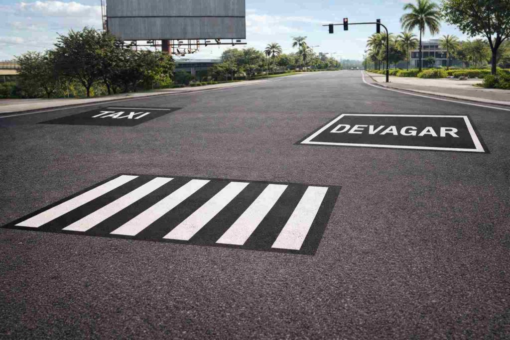 Imagem realista de uma via urbana moderna apresentando exemplos de sinalização horizontal preta conforme utilizada no padrão brasileiro para reforço de contraste e acabamento visual em marcações viárias, aplicada sobre pavimento claro com demarcações de orientação e separação de áreas, ambientada em cenário urbano com iluminação natural suave e composição educativa institucional.