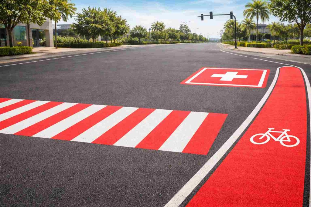 Imagem realista de uma via urbana moderna exibindo exemplos de sinalização horizontal vermelha conforme utilizada no trânsito brasileiro, com faixa de pedestres vermelha e branca destacada, área de circulação exclusiva para ciclovia/ciclofaixa em vermelho e marcação de área de emergência/saúde sinalizada sobre o asfalto, ambientada em cenário urbano com iluminação natural suave e composição educativa institucional.